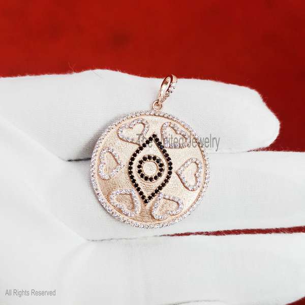 "A stunning fusion of love and protection, this large round medallion features a textured rose gold-tone finish. It is intricately designed with a central black-stone evil eye surrounded by a halo of shimmering hearts and a brilliant pave-set border. Product Details Material: High-quality 925 Sterling Silver. Design: Circular medallion with a protective evil eye center and six pave-lined hearts. Accent Details: Contrasting black micro-pave stones for the eye and clear sparkling stones for the hearts, border, and bail. Texture: Unique brushed/satin ""stardust"" finish on the base metal for added depth. Finish: Luxurious Rose Gold plating over sterling silver. Style: Symbolic Statement / Talisman / Modern Romantic."