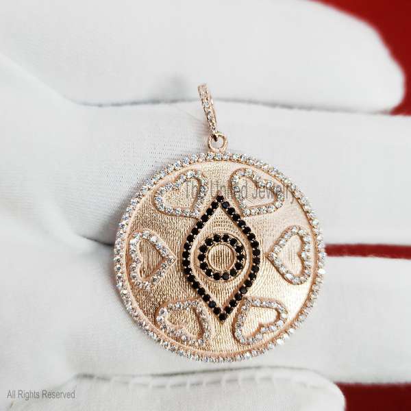 "A stunning fusion of love and protection, this large round medallion features a textured rose gold-tone finish. It is intricately designed with a central black-stone evil eye surrounded by a halo of shimmering hearts and a brilliant pave-set border. Product Details Material: High-quality 925 Sterling Silver. Design: Circular medallion with a protective evil eye center and six pave-lined hearts. Accent Details: Contrasting black micro-pave stones for the eye and clear sparkling stones for the hearts, border, and bail. Texture: Unique brushed/satin ""stardust"" finish on the base metal for added depth. Finish: Luxurious Rose Gold plating over sterling silver. Style: Symbolic Statement / Talisman / Modern Romantic."